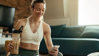 Happy athletic woman using mobile phone while drinking a protein shake at home