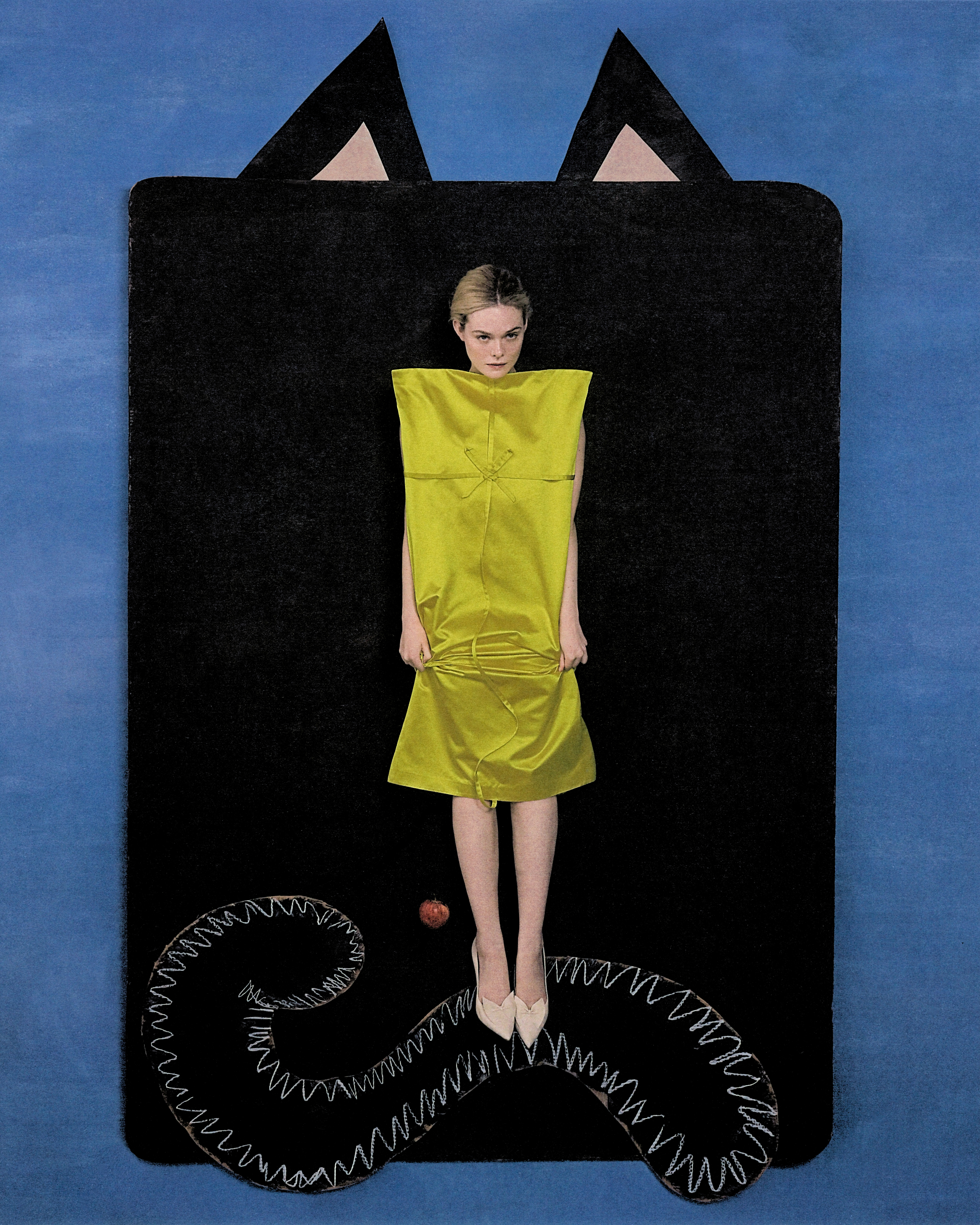 Elle Fanning shot by Szilveszter Mak&amp;oacute;. She is lying on a large black cardboard surface with cat ears and tail. She is wearing a yellow silk dress with with square neckline and bow detailing.