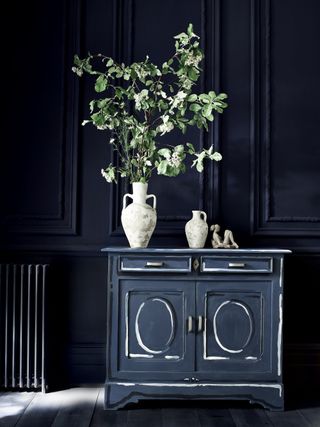 dark blue apartment living room with painted cabinet, vase with foliage and ornaments on top, painted wooden floor, painted radiator, panelled walls