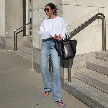 female fashion influencer Sasha Mei poses outside a city building with a low bun, aviator sunglasses, a long-sleeve white sweatshirt, baggy straight-leg jeans, a black top-handle bag, and blue and black contrast flip-flop kitten heels