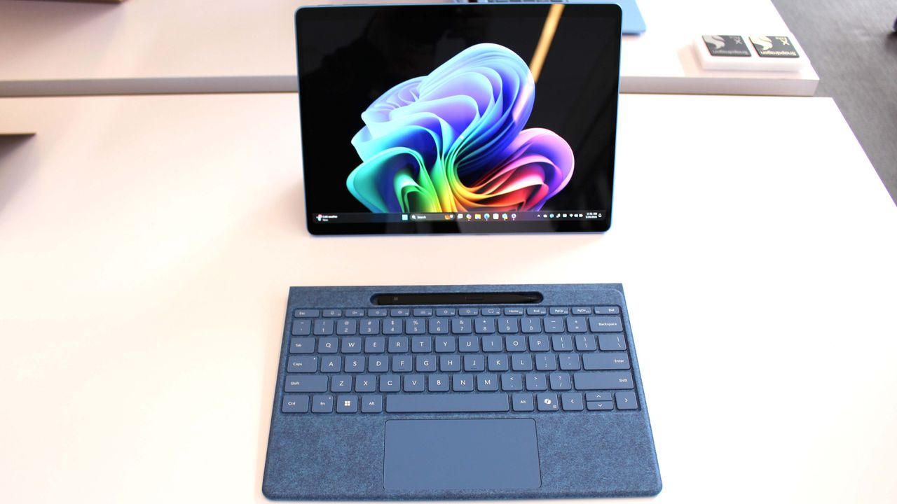 A sleek Surface Pro 11 tablet with a vibrant, colorful swirl on its screen is connected to a detachable blue keyboard, placed on a light wooden desk.