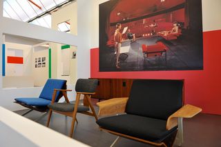 Three different style chairs sit in front of a large advertising board in the exhibition space. The ad. board features people sat in different chairs in an auditorium-like room. .