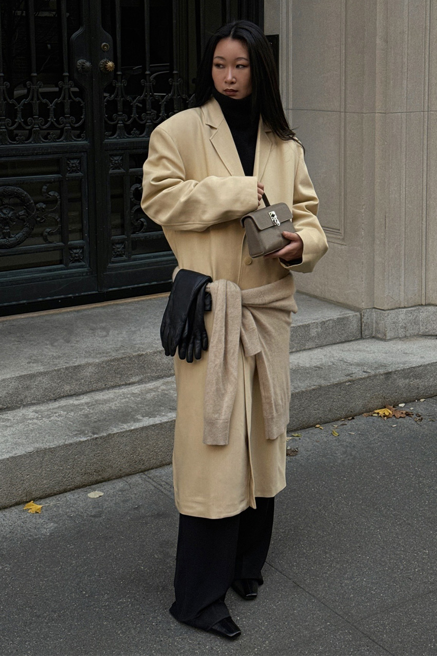 West Village winter trends are shown in a street-style photo of a woman standing in front of a black doorway in a concrete building, wearing her winter outfit. She's wearing a black turtleneck sweater and black wide-leg trousers layered under a cream coat with a matching cardigan tied around the waist and black gloves layered over the cardigan styled with black square-toe ankle boots and a taupe clutch bag.