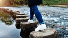 A woman walks across stepping stones in a river, only her legs showing.