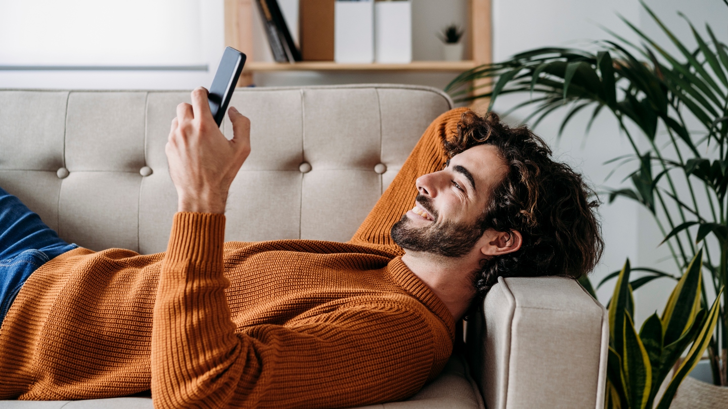 Man lying on couch holding phone and smiling at it