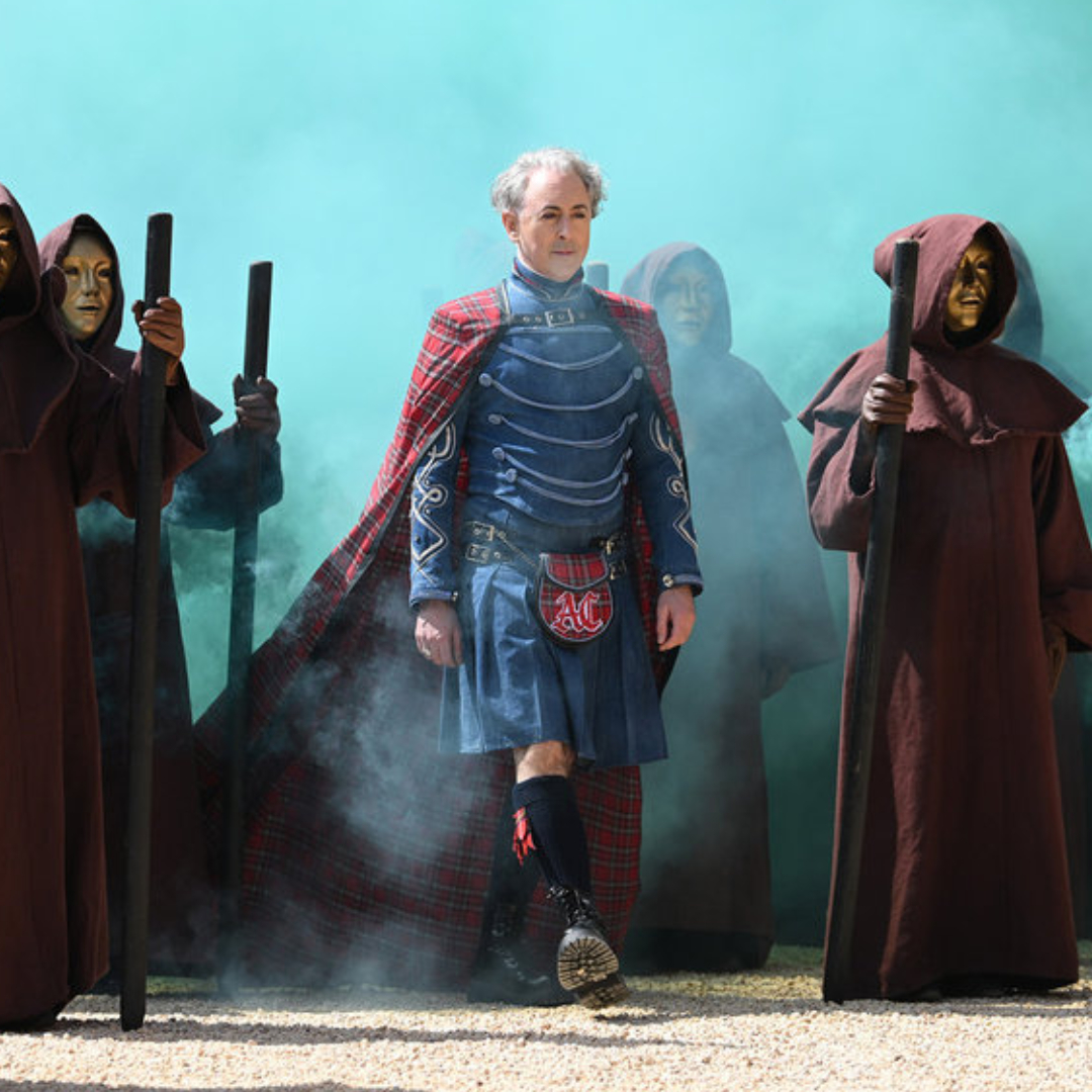 Host Alan Cumming wears armor and a cape while standing amid cloaked figures with gold masks in a cloud of blue smoke, in NBC's 'The Traitors: New Blood.'