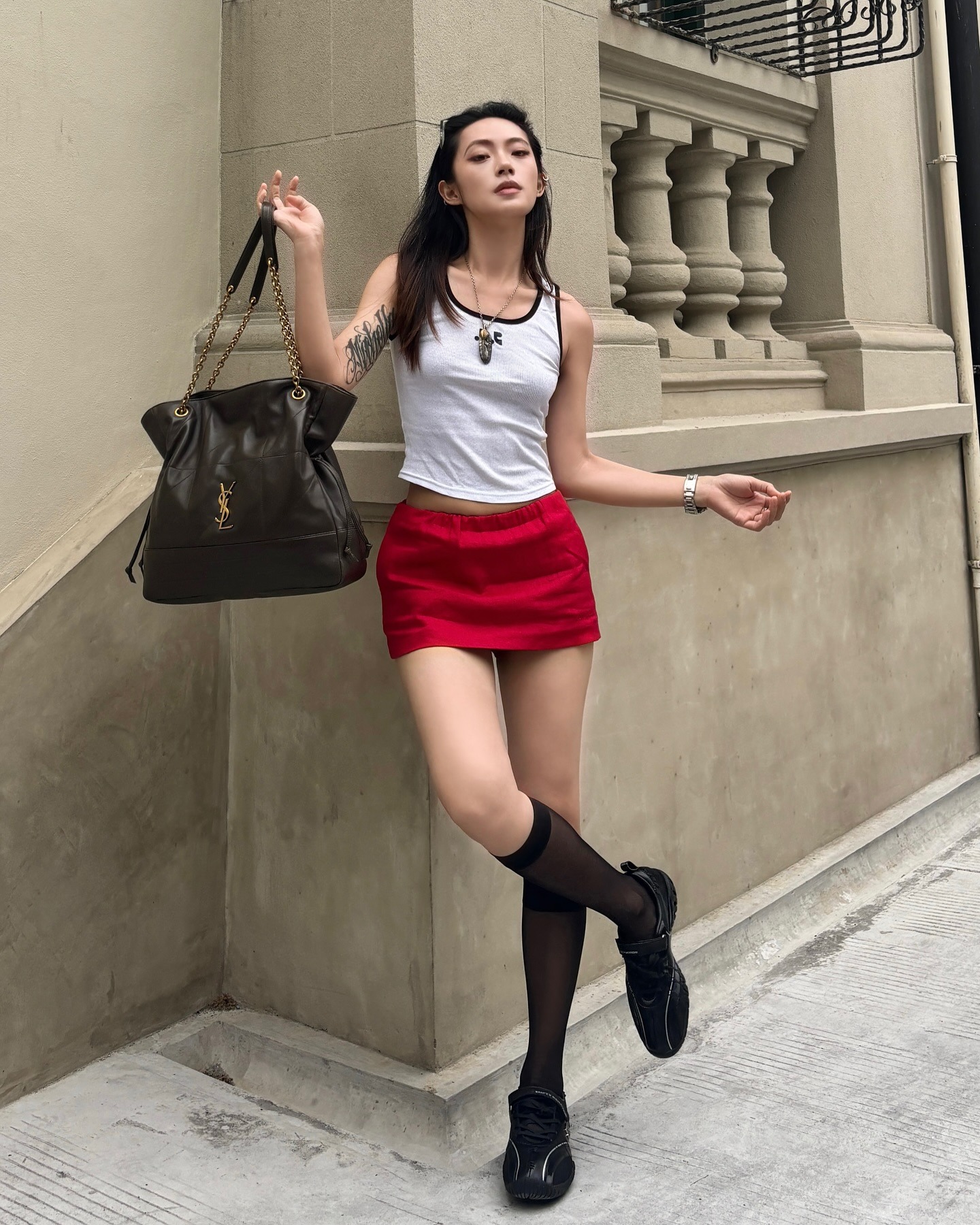 A woman wearing a white tank top, red skirt, black shoes, and holding a black leather YSL bag