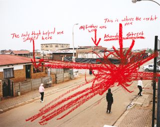 Color photograph of people stood on a street. The photo is marked with red annotations saying: “The lady that helped me stay here”, “My father was here”, “This is where the car hit me” and “Khumalo Street.”