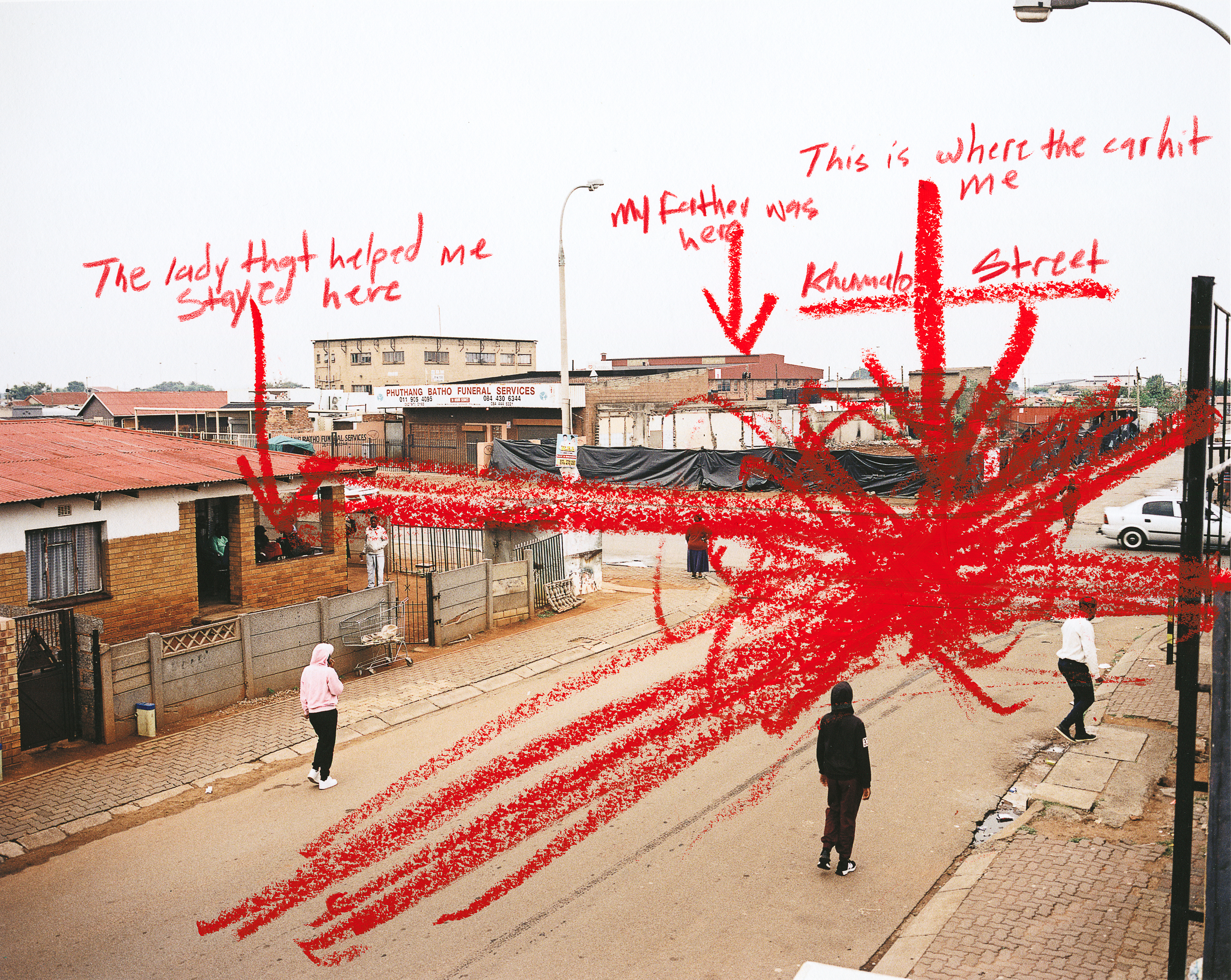 Color photograph of people stood on a street. The photo is marked with red annotations saying: “The lady that helped me stay here”, “My father was here”, “This is where the car hit me” and “Khumalo Street.”