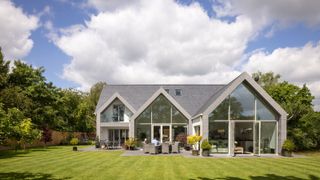 large modern house set in gardens with expanse of green lawn, 3 pitched roof gable ends along rear of property with grey frames