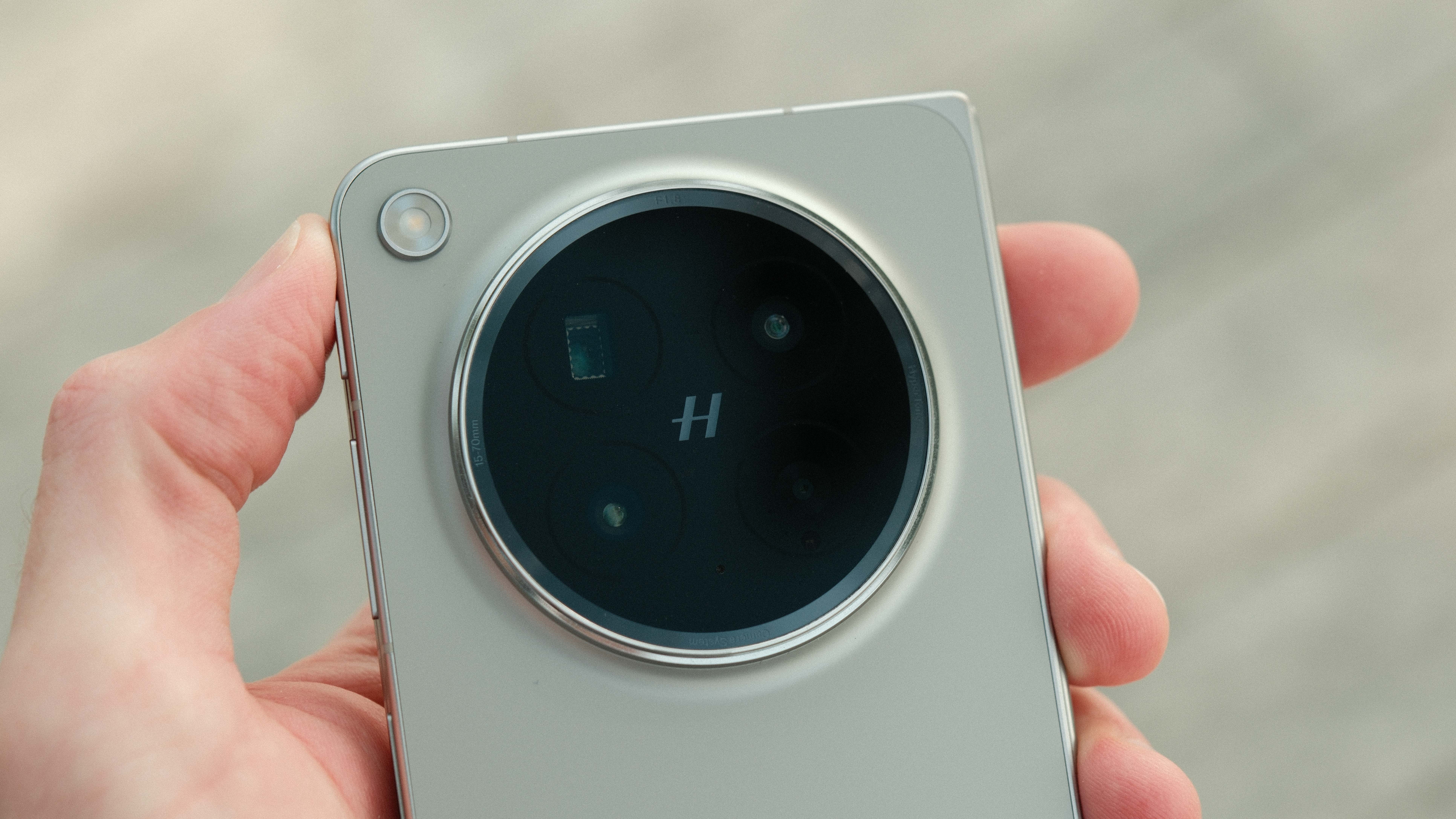 A person holding the phone, providing a clear view of the rear design. Text etched around the camera bezel indicates a focal range of "15-70mm" and an "f/1.8" aperture.