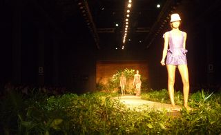 Model in a lilac outfit and hat, on a runway lined with foliage