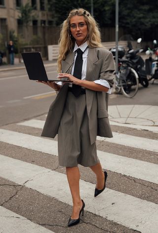 Milan Fashion Week spring 2025 street style photo of a woman wearing geek chic trend in the form of a gray blazer and matching skirt with a white button-down shirt and black tie and black pumps