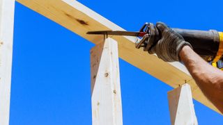 Gloved hand holding reciprocating saw cutting nails in a timber frame against a bright blue sky