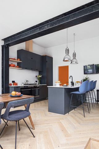 Black sleek kitchen with island. Wood chevron flooring, blue bar stools and glass pendant lights over island