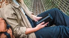 PINE Beat bluetooth speaker in the hands of a woman wearing a beige coat, white short and blue trousers. She appears to be sitting in a rope bridge