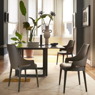 small elegant dining room with modern brown chairs, square table, rug, plants, vases, artwork, brown walls, large window in background