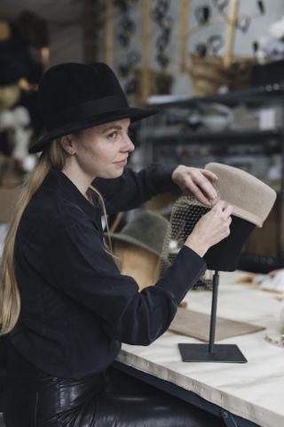 A portrait of hatmaker Gigi Burris putting a veil on one of her hats