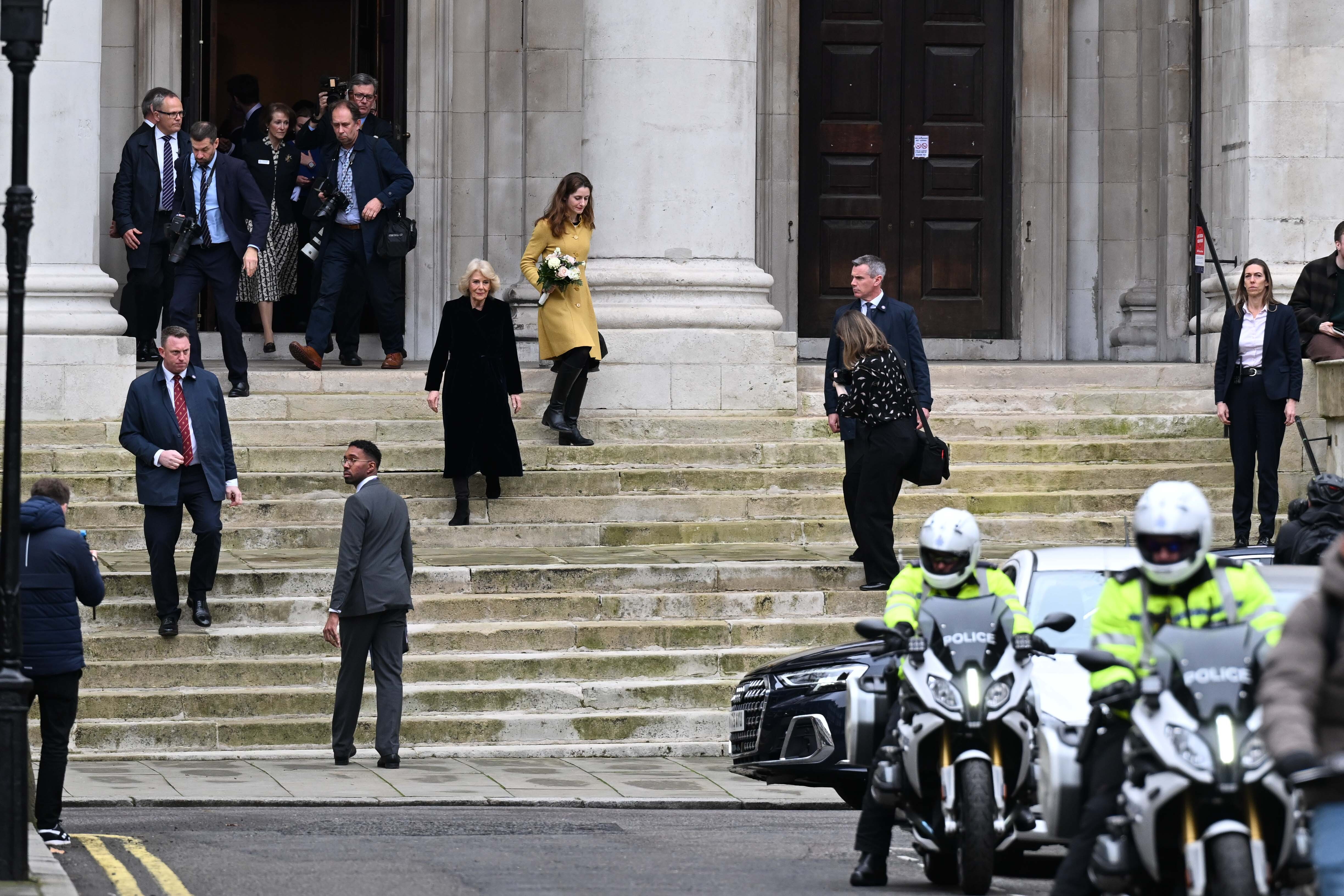 Queen Camilla walking down the stairs of a building surrounded by police on bicycles