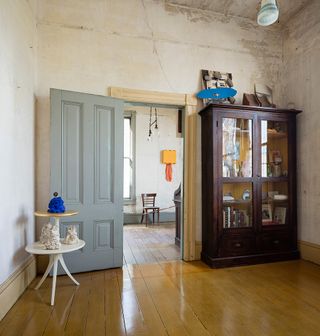 A view of the guestroom. Light-colored wooden floors, with a dark wooden cabinet with glass doors standing against a wall. The walls are worn out, light cream color.