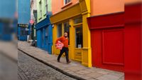 A man wearing a bright orange sweater and cap walks down a cobblestone sidewalk carrying a red box past a row of vibrant, colorful storefronts.