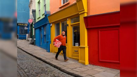 A man wearing a bright orange sweater and cap walks down a cobblestone sidewalk carrying a red box past a row of vibrant, colorful storefronts.