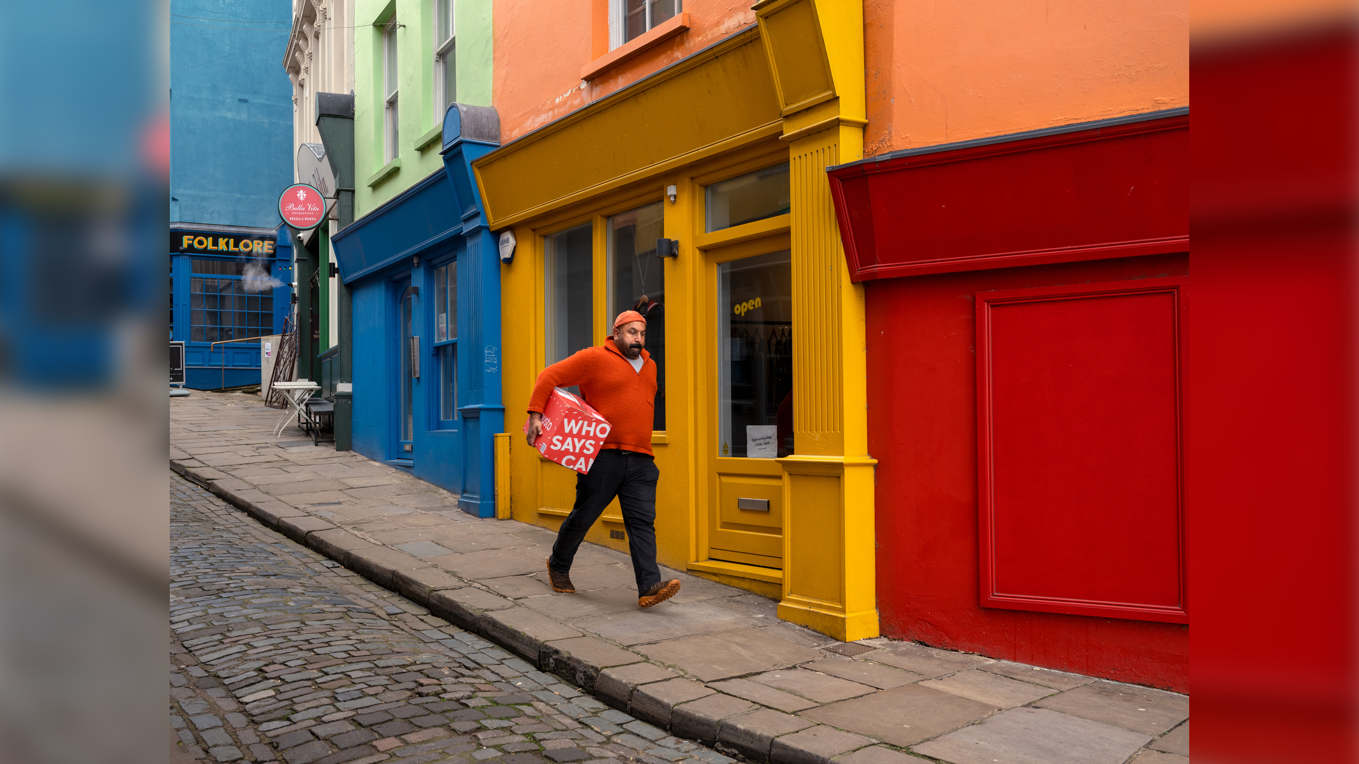 A man wearing a bright orange sweater and cap walks down a cobblestone sidewalk carrying a red box past a row of vibrant, colorful storefronts.
