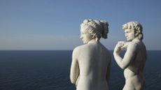 Two Roman statues of a man and a woman with the ocean in the background