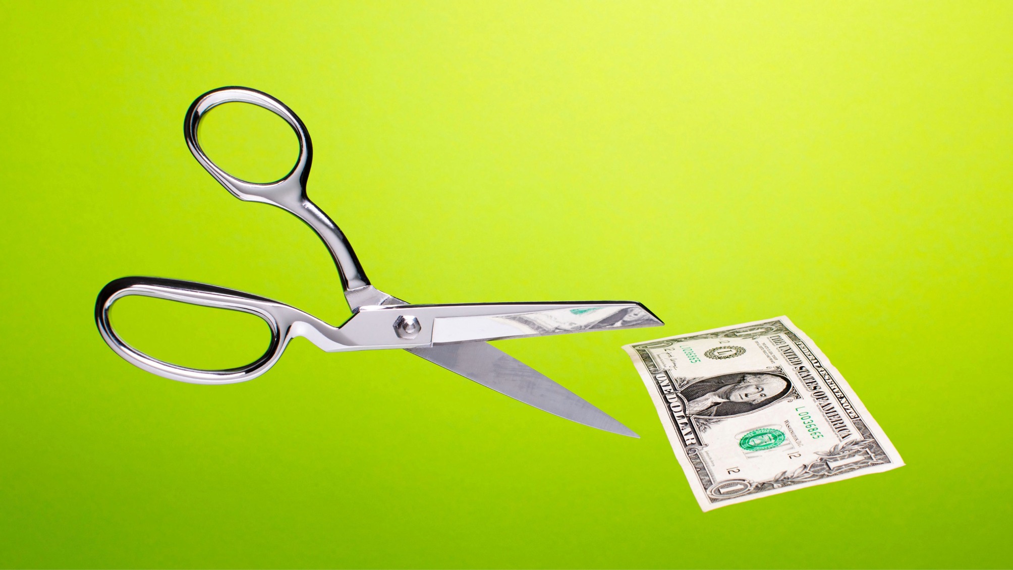 Stainless steel scissors in mid air about to cut US $1 dollar bill over gradated green background