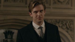 Dan Stevens smiles wryly in front of a fireplace in Downton Abbey.