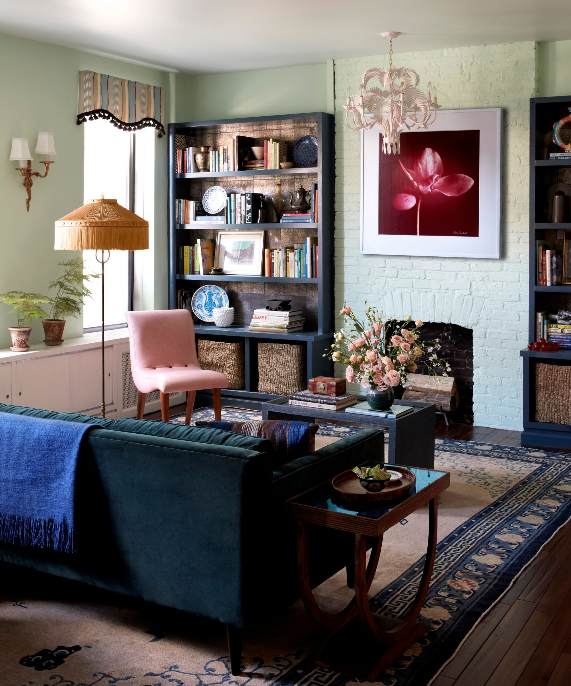 A green painted fireplace in an eclectic, colorful living room with a large patterned rug, velvet sofa, statement artwork and dark blue bookshelves with a white chandelier.