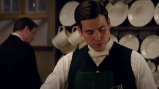 Rob James-Collier works away while Brendan Coyle stands behind him listening in Downton Abbey.