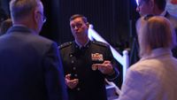 a man in a black military uniform speaks to a group of attendees on a conference ballroom floor