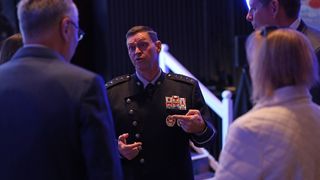 a man in a black military uniform speaks to a group of attendees on a conference ballroom floor