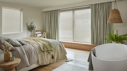 A large bedroom with sage green curtain window treatment decor, white blinds, large bed and white bathtub