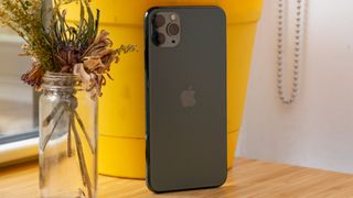 Hvordan kommer iPhone Flip mon til at se ud? Billedet viser bagsiden af en iPhone 11 Pro Max, som står på et træbord op ad en gul potte ved siden af en vase med tørrede blomster