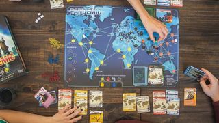 best board games for two players - a hand reaching onto a pandemic board, cards lined up at its edge