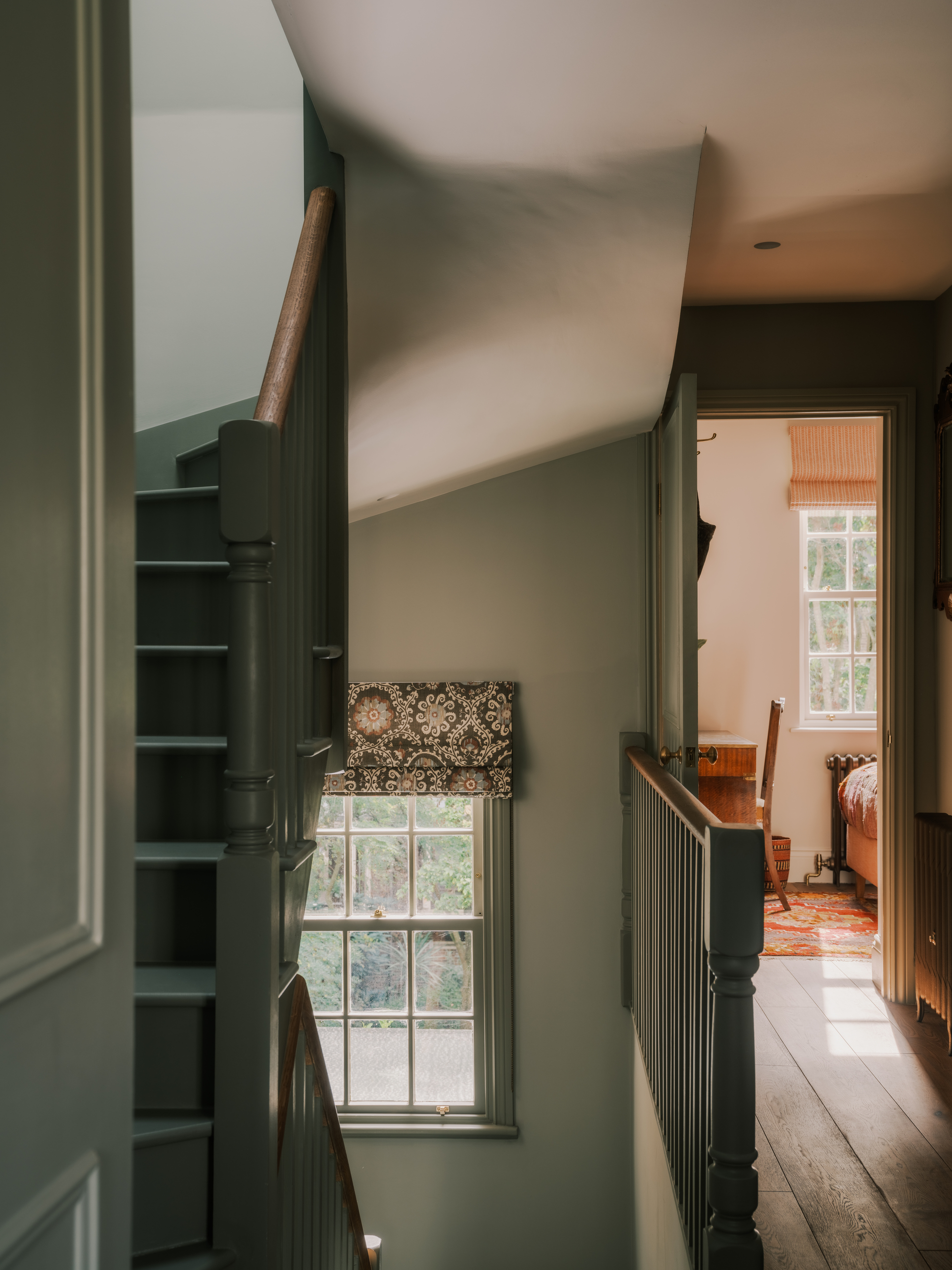 Image of a sage green hallway with a window that has a patterned roman blind. There is a door open upstairs that shows a pink bedroom.