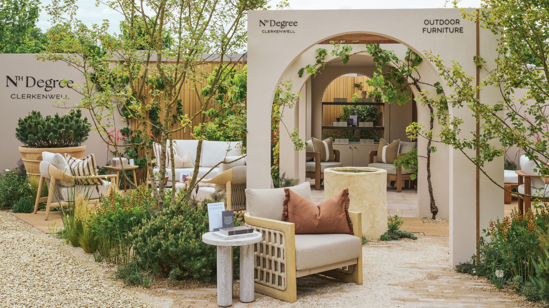 An outdoor living room with wood armchairs, colored cushions, a gray marble side table, arched doorways, terracotta pots, and a water pot feature