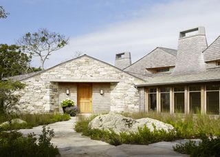 Front walkway ideas illustrated by two pathways surrounded by tall grasses leading to a stone gray house.
