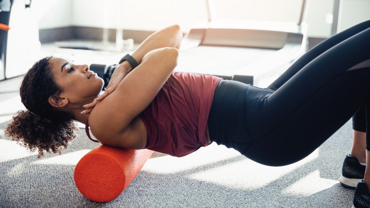 A woman wearing sportswear stretches her back on a foam roller; her feet are planted on the floor, knees bent, arms crossed over her chest, and the foam roller is propped under her shoulders.