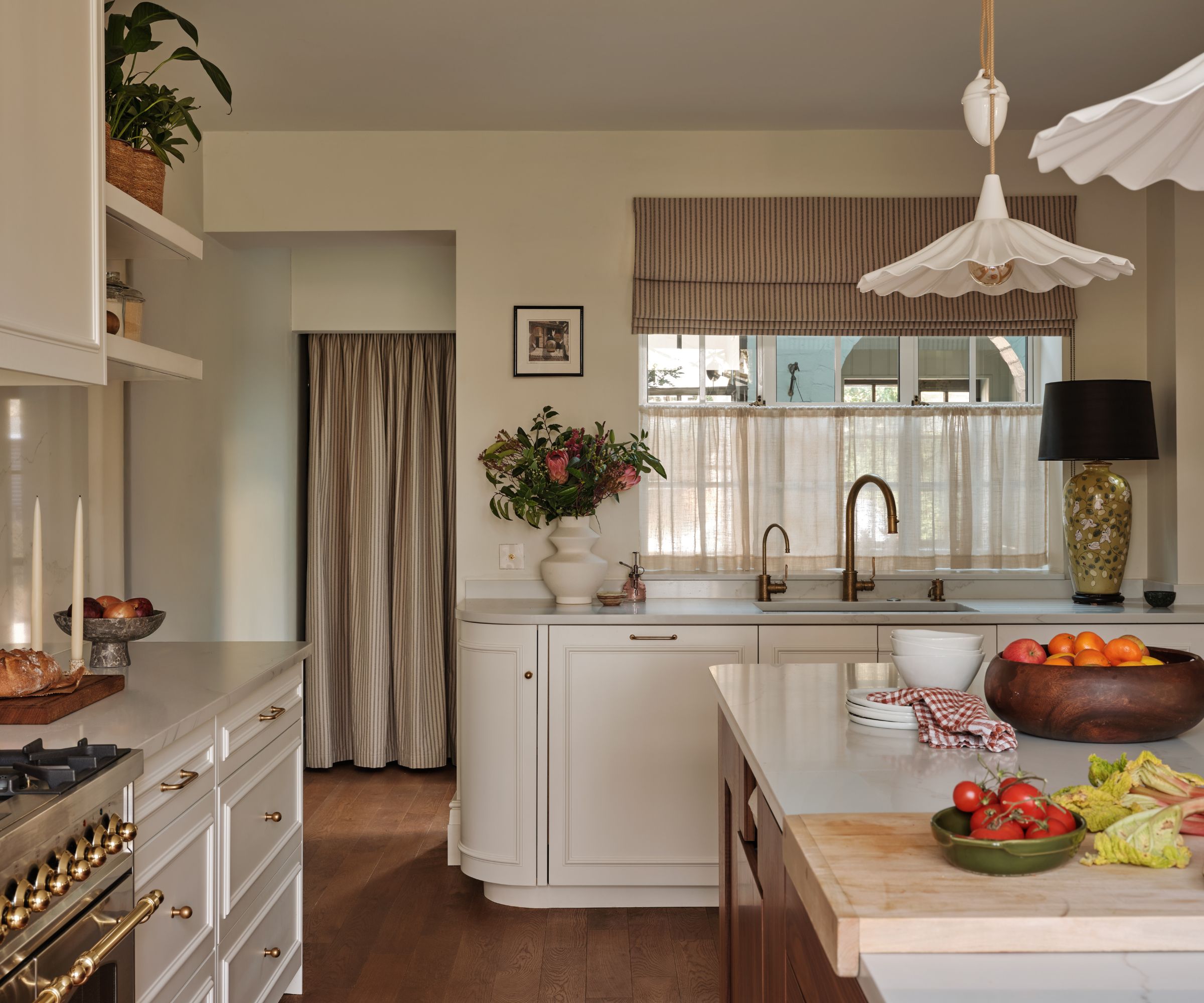 Warm, traditional kitchen with white cabinetry, brass faucet fixtures, a center island with fresh produce, and striped Roman shades on the window