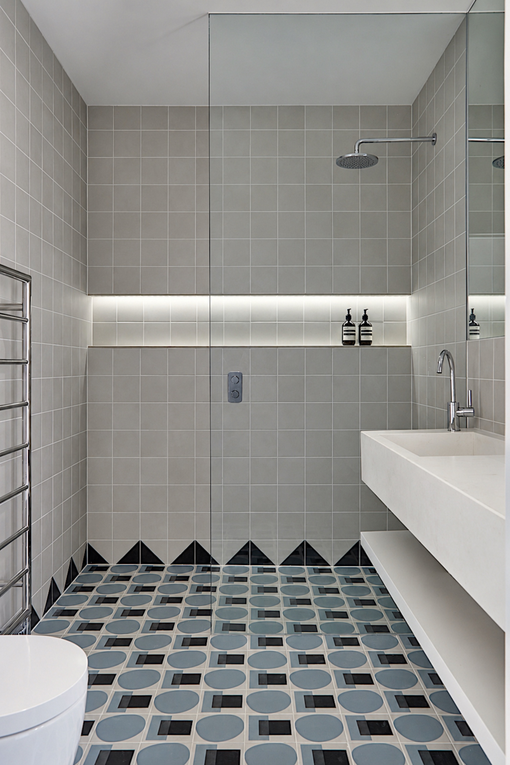 Bathroom with gray square wall tiles, a black triangular tile border, blue geometric floor tiles, a glass shower screen and a white floating vanity.