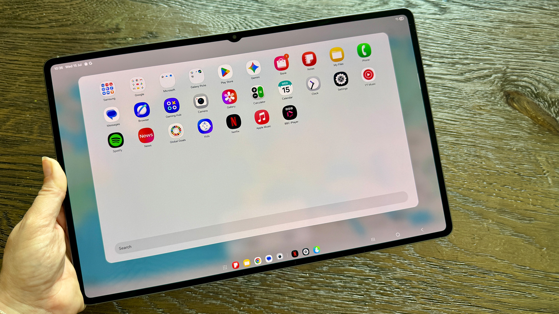 Samsung Galaxy Tab S11 Ultra tablet held in hand above wooden table, on screen is app folder