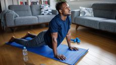 A man performs a yoga cobra pose in a living room on a mat. His hands are planted on the mat, arms straight, and his torso is upright as his legs lie on the ground behind him. In the background is a pair of couches, a bookcase and two light dumbbells.