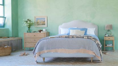 Refreshing and calm bedroom with light mint walls, lived-in bedlinen layers, and rustic wooden furniture, with shapely upholstered headboard.