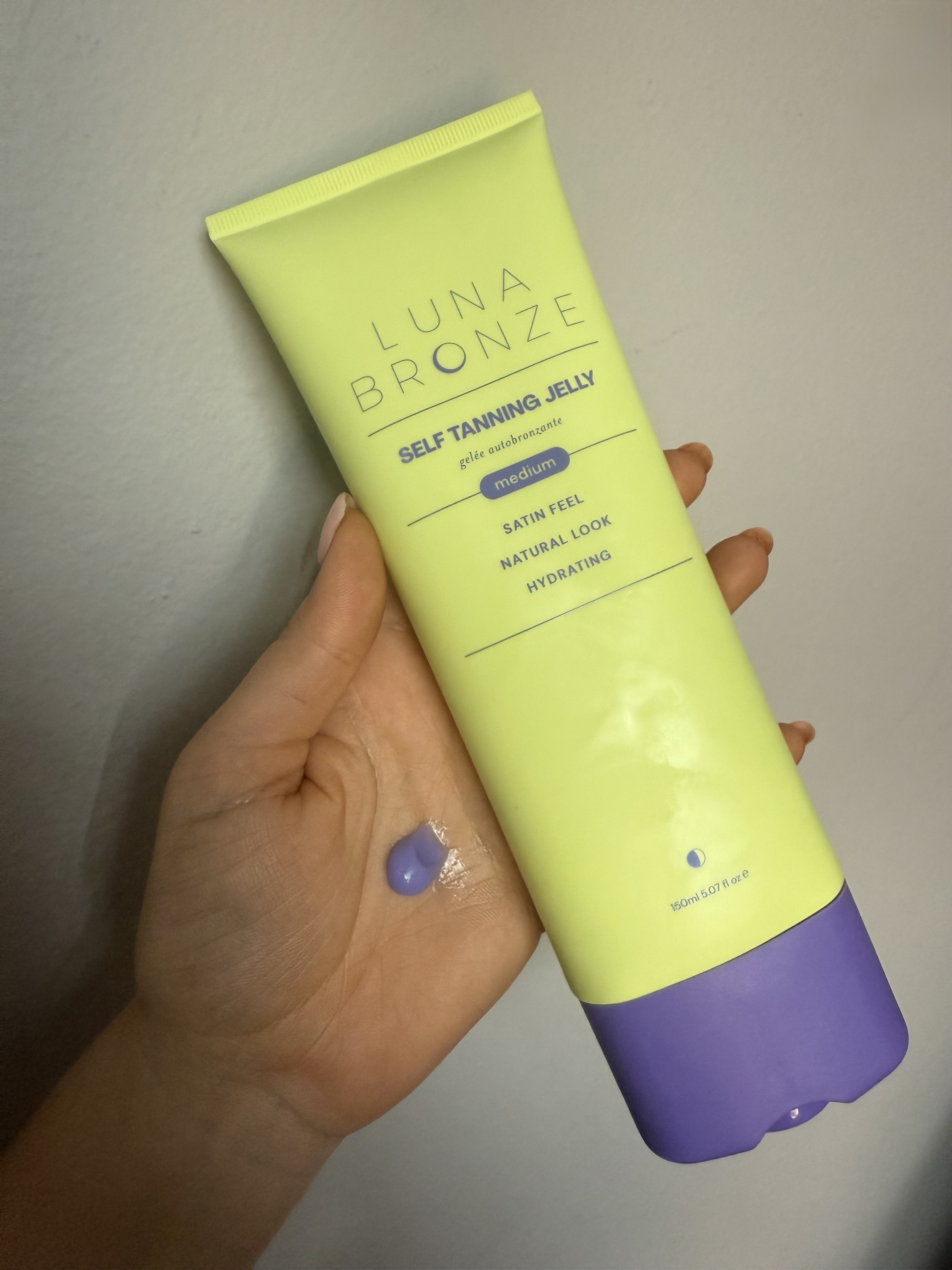 Brooke knappenberger holding up bottle of luna bronze self tanning jelly against grey wall