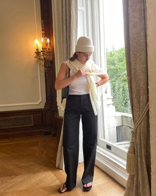 Anna Laplaca at a Bevza event during NYFW wearing a cream Gigi Burris hat with a matching scarf, a white tank top, black trousers, and black flip flops.