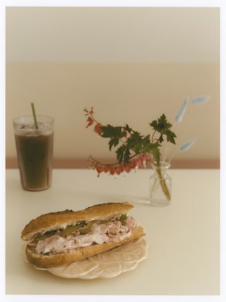 A ham and gherkins sandwich sits atop a white table in front of an iced coffee glass and a transparent glass jar filled with pink and blue dried flowers, all of which stand against a rose wall.