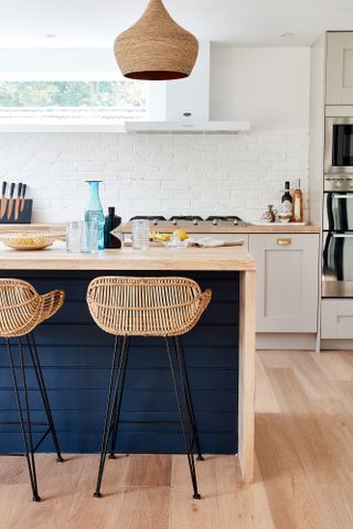 How to use exposed brick: white painted brick wall in background of phoeo with white kitchen, dark blue island and rattan pendant and bar stools